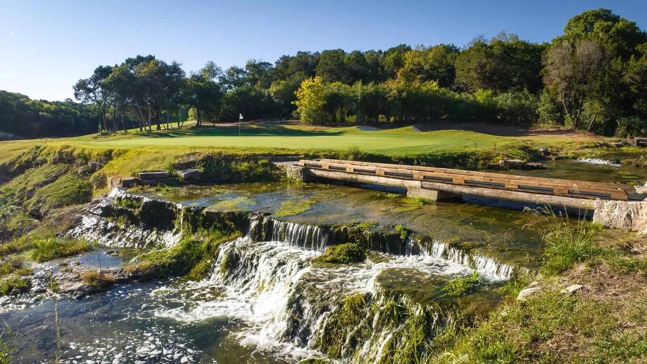Scenic golf green beside a cascading stream and wooden bridge at a Texas Hill Country course.